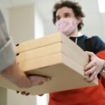 Delivery worker handing over pizzas while wearing a protective mask indoors, illustrating safe delivery practices during a pandemic.